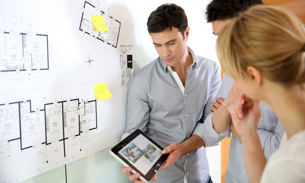 Man showing a proposed design in an iPad to a couple with a floorplan haing in the wall